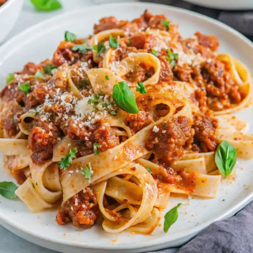 A white plate holds wide fettuccine pasta generously coated in rich, meaty Lamb Bolognese sauce. It is garnished with grated Parmesan cheese and fresh chopped parsley, ready to serve.