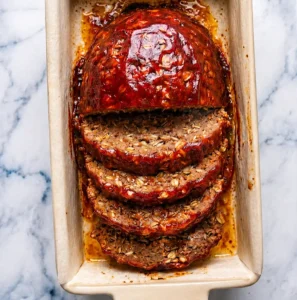 sliced homemade meatloaf with oatmeal, covered in a glossy tomato glaze, arranged inside a ceramic loaf pan on a marble background.