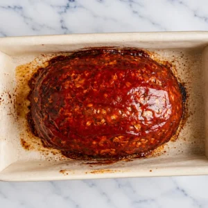 baked homemade meatloaf with oatmeal, topped with a glossy, sweet tomato glaze, resting in a rectangular ceramic baking dish on a marble countertop.