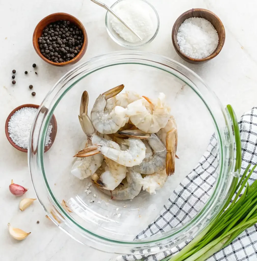Ingredients for Salt and Pepper Shrimp glass bowl of raw, and shrimp surrounded by. These ingredients include bowls of black peppercorns, coarse salt, a cornstarch, garlic cloves, and fresh green onions.