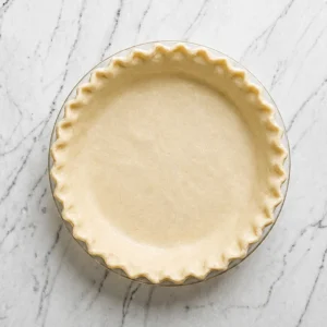 A raw pie with fluted edges, placed inside a glass pie dish and set on a white marble surface, ready to be filled with sausage quiche filling.