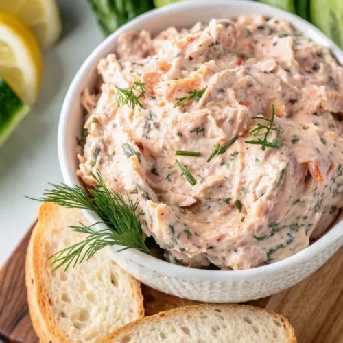 finished Smoked Trout Dip served in a white bowl, garnished with fresh dill. The creamy, pink dip is presented on a wooden board with slices of toasted baguette bread and garnishes like lemon and cucumber in the background, ready for serving.