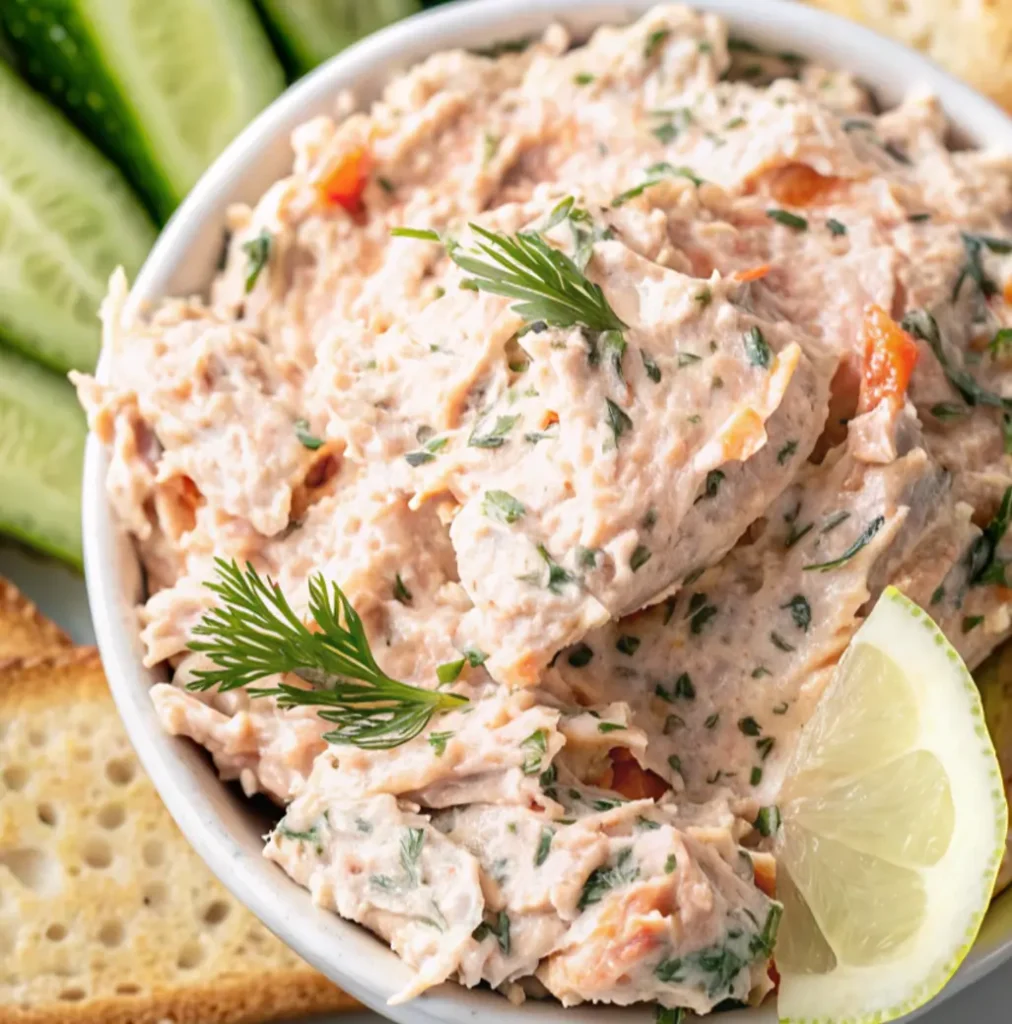 finished Smoked Trout Dip served in a white bowl, garnished with fresh dill. The creamy, pink dip is presented on a wooden board with slices of toasted baguette bread and garnishes like lemon and cucumber in the background, ready for serving.