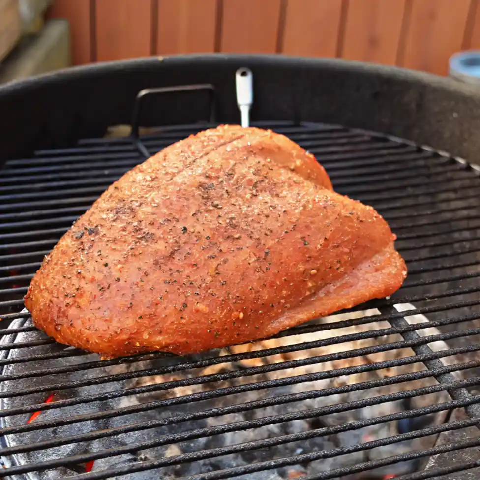 A large, seasoned turkey breast with a reddish-orange spice rub cooking on a black circular grill grate over hot coals, with a meat thermometer inserted."