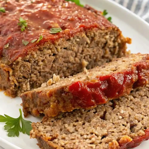 A close-up of a sliced, meatloaf recipe with oatmeal on a white platter. The moist interior texture shows the oat binder, and the top is covered with a thick, glossy tomato glaze garnished with fresh thyme and parsley.
