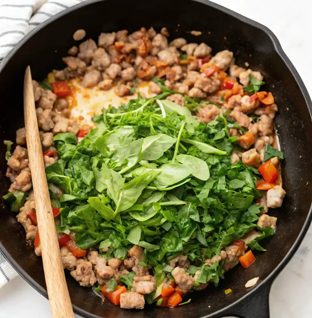 A wooden spoon rests beside crumbled sausage and red pepper mixed with fresh green leafy vegetables in a dark cast iron pan for a quiche filling.