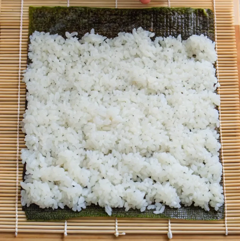 A rectangular sheet of dark nori seaweed rests on a bamboo sushi rolling mat. A layer of fluffy, white sushi rice is evenly spread across almost the entire surface of the nori