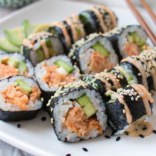 A large white plate holds several slices of Spicy Shrimp Roll cut into half-inch pieces. The filling consists of small pieces of spicy orange shrimp, green cucumber, and avocado. Some rolls are topped with spicy mayonnaise sauce and sesame seeds, with chopsticks placed in the corner.
