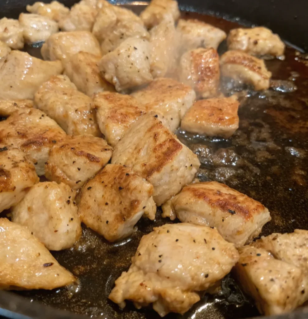 A close-up of seasoned chicken pieces cooking in a sizzling black skillet, showing light browning and steam rising, indicating the grilling or frying step.