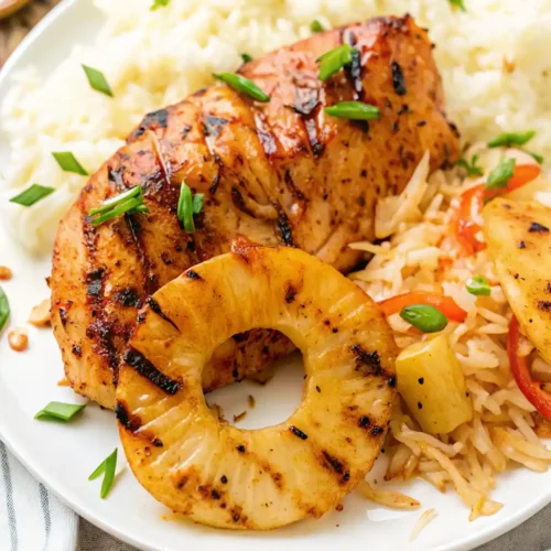 This plate features a serving of Hawaiian BBQ Chicken Recipe, showing a glossy, grilled chicken breast and a grilled pineapple ring. It's served with a scoop of fluffy white rice and a portion of pineapple fried rice, all garnished with chopped scallions.
