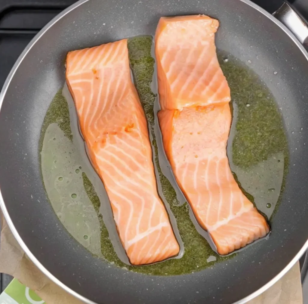 Two fillets of raw salmon, skin-side down, are searing in a black non-stick pan containing a generous amount of oil