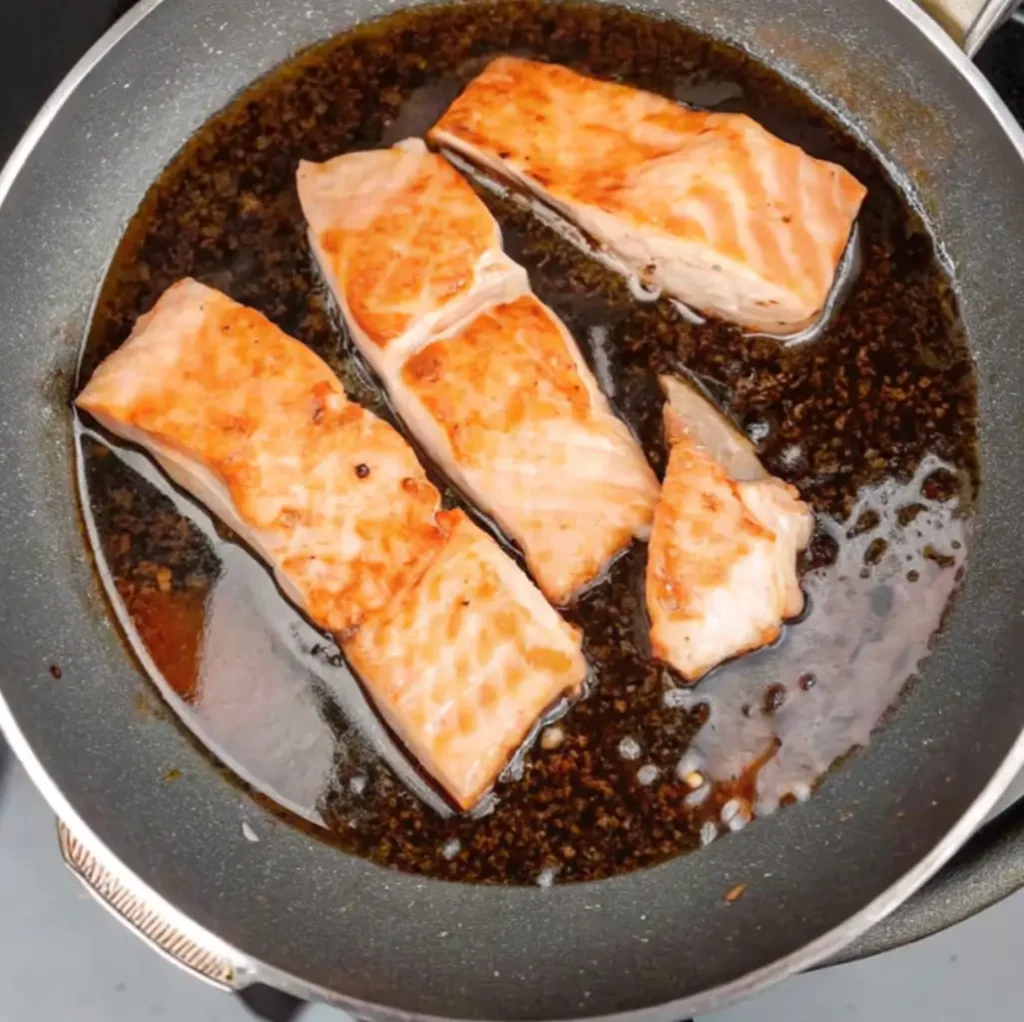 Four pieces of seared salmon fillet are now simmering in a rich, dark brown sauce within a frying pan