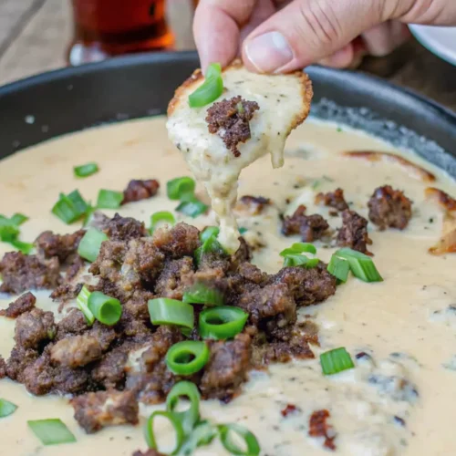 A hand dips a crispy toast round into the creamy, tan-colored hot sausage beer cheese dip, pulling up a spoonful of melted cheese and crumbled sausage garnished with green onions.