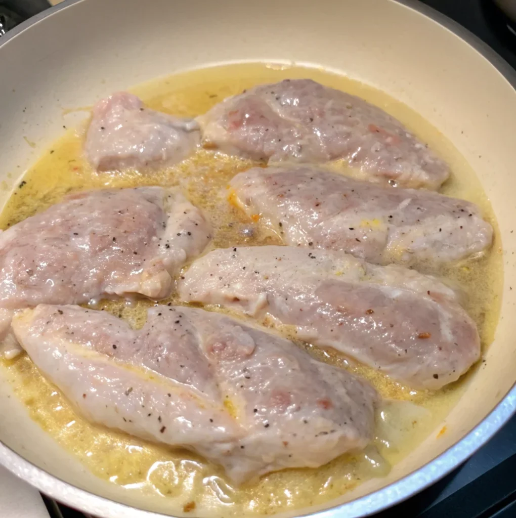 Five pieces of seasoned, raw chicken are cooking in a sizzling butter or oil mixture in a non-stick skillet. They are lightly coated and showing early signs of browning.