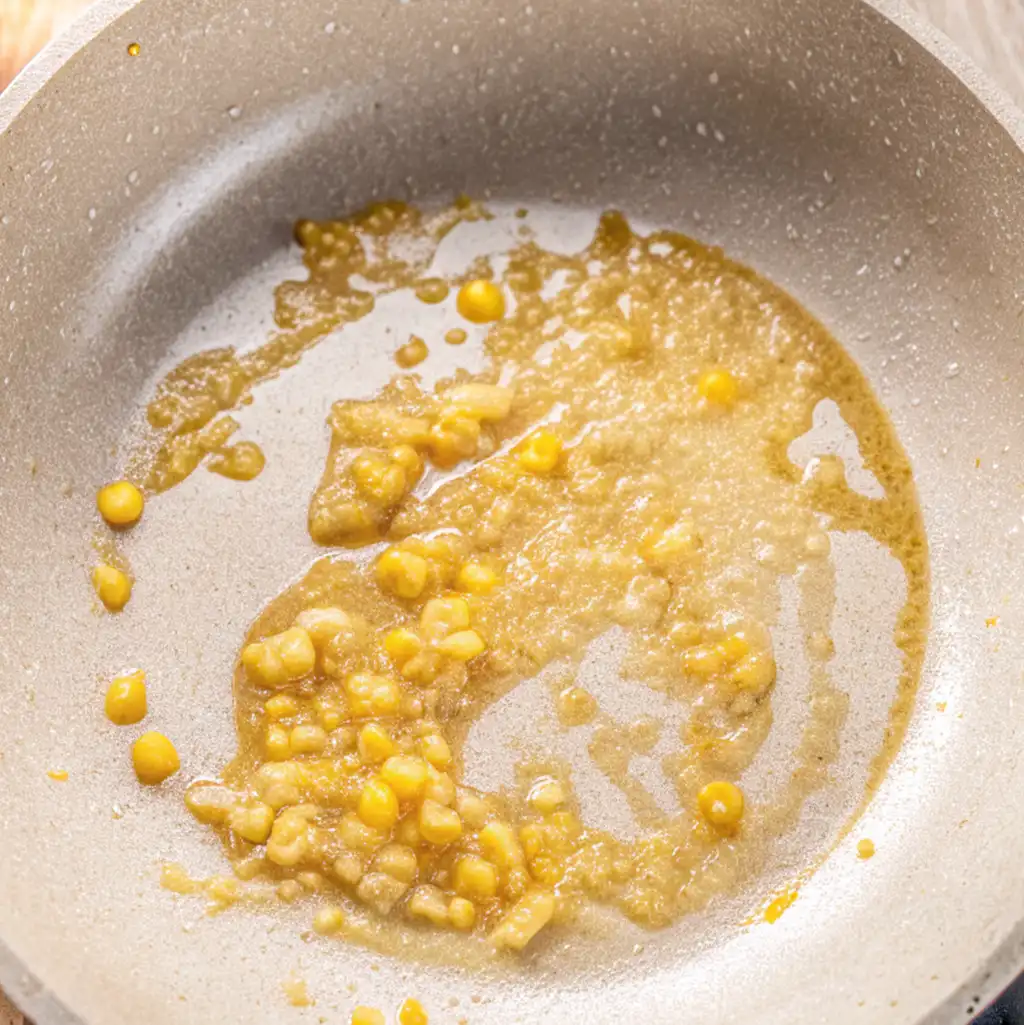 The light-colored pan shows garlic and ginger fried in a golden, oily, slightly caramelized mixture, as part of the preparation of seafood ramen.