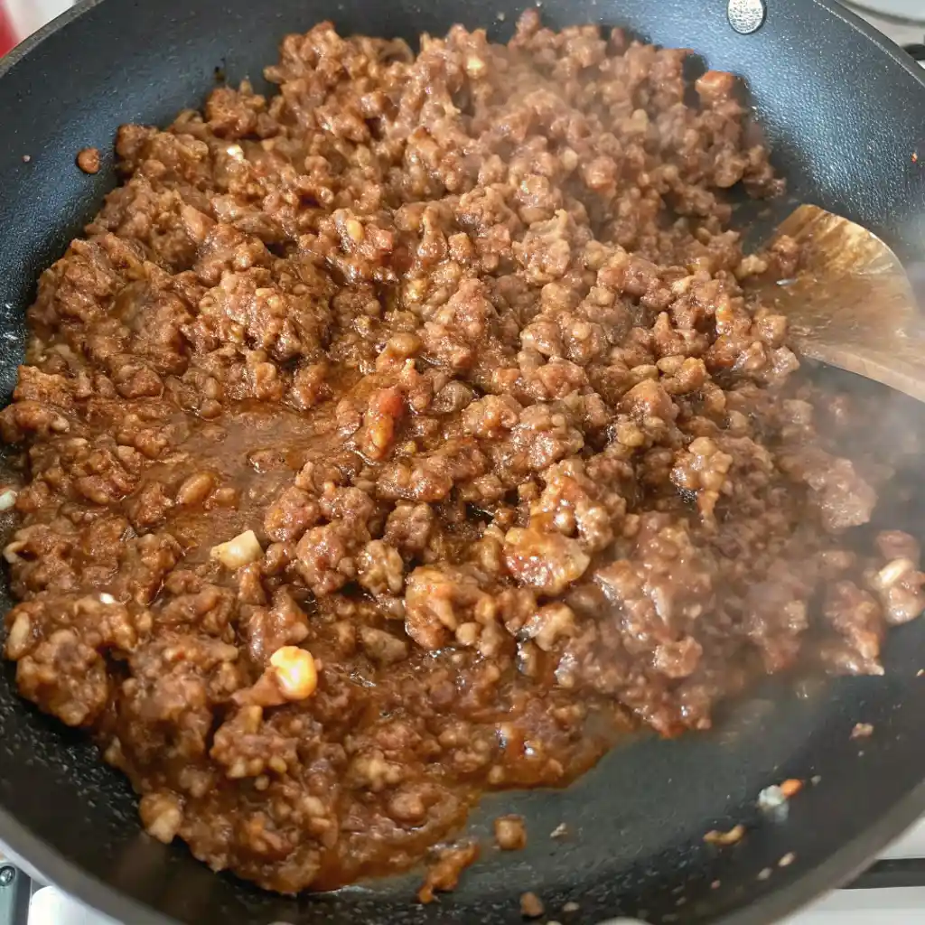 Sloppy Joe filling simmering in a black non-stick skillet on a stovetop. The rich, thick, brown ground meat mixture is being stirred with a wooden spoon.