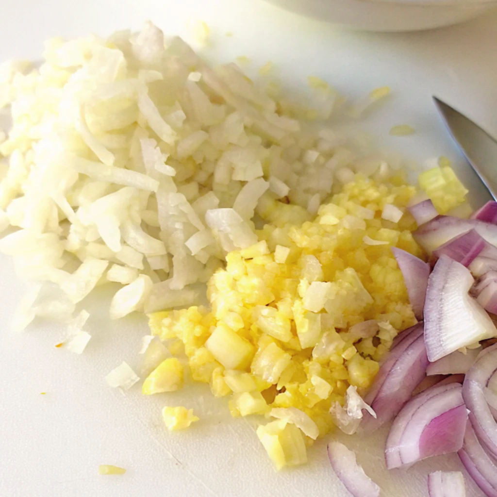 three piles of diced and chopped ingredients: white onions, yellow minced garlic, and sliced red onions, with a knife tip visible on the right.
