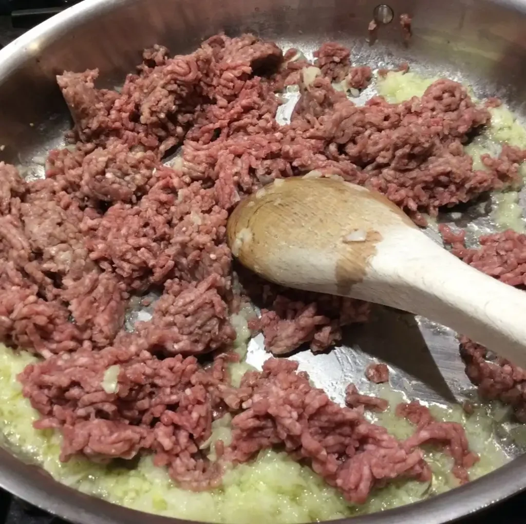 Close-up of pink raw minced Wagyu beef being added to a stainless steel pan with diced and sautéed onions, in the center of a large wooden spoon for mixing.