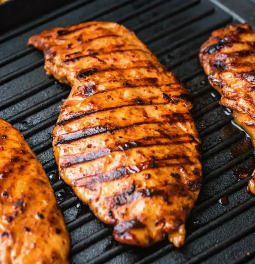 This is a close-up of a marinated chicken breast grilling on a hot black grate. The chicken has a rich, orange-brown BBQ glaze and distinct dark grill marks running across its surface. for Hawaiian BBQ Chicken Recipe