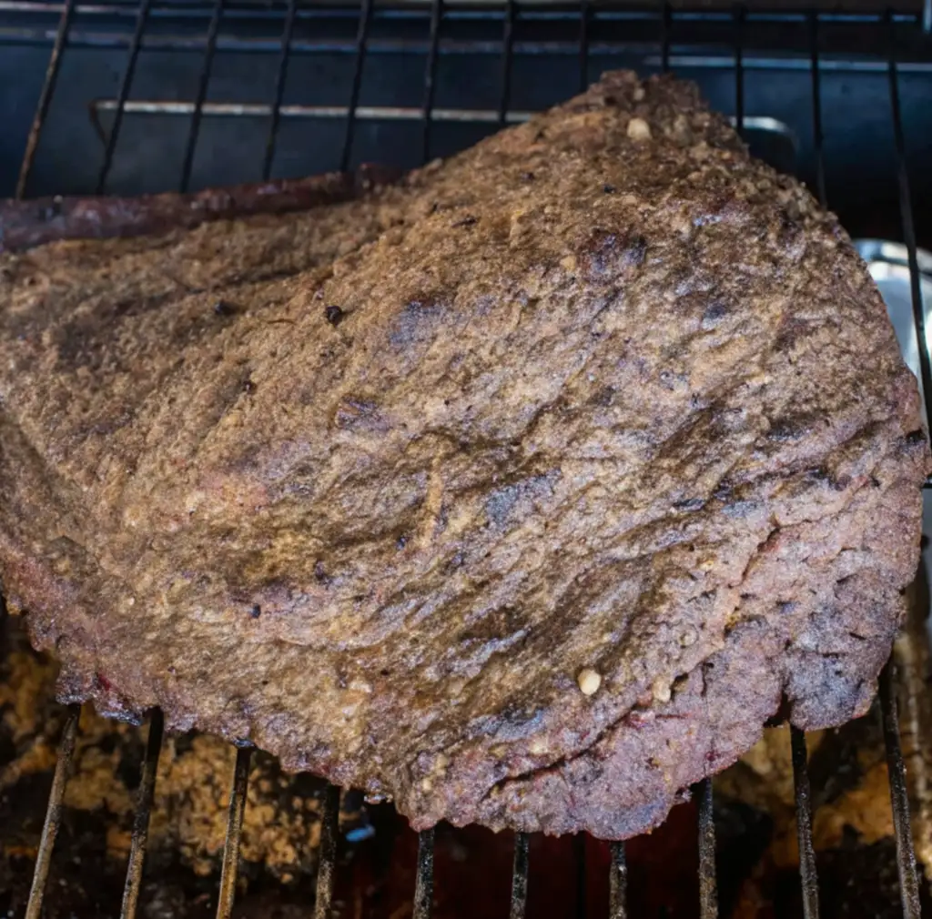 A large, thick piece of smoked corned beef brisket rests on a wire rack over a grill, showcasing a deeply crusted, savory bark. The rich, dark brown exterior suggests hours of slow cooking.
