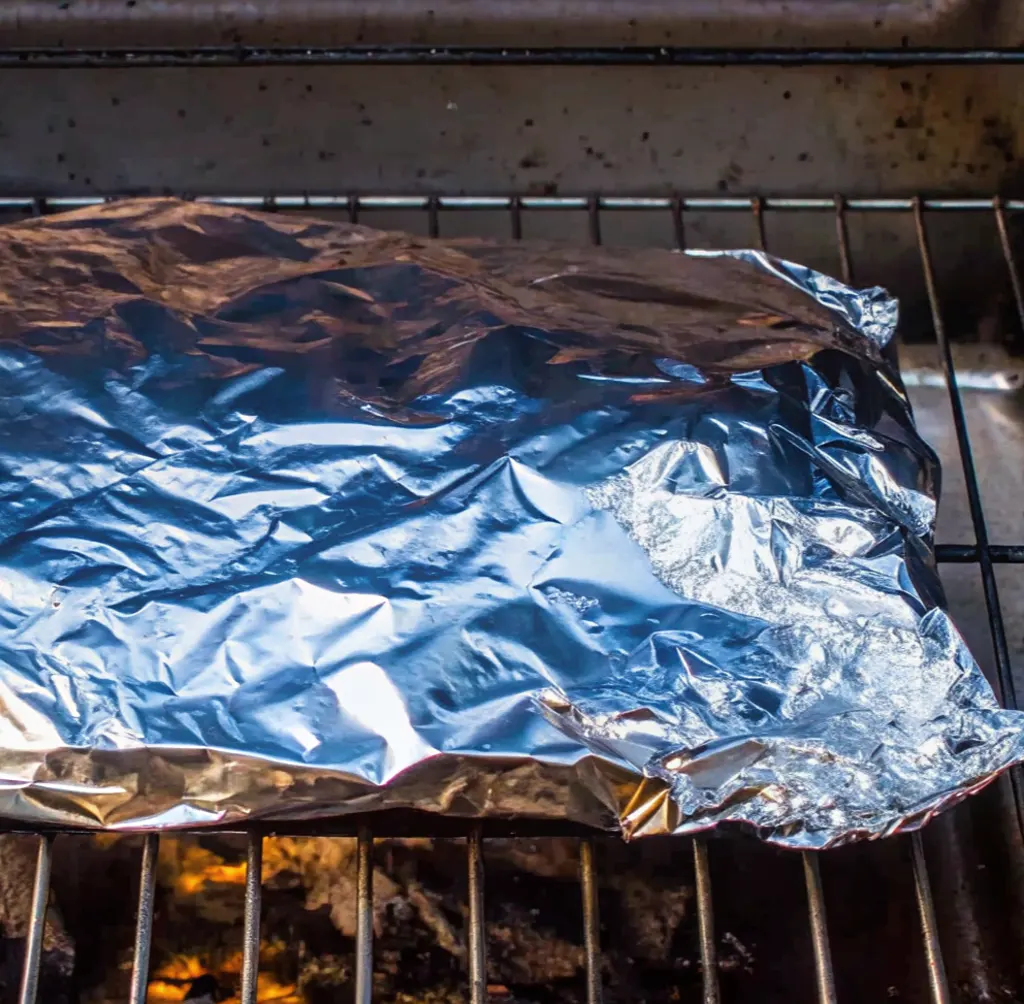 A piece of smoked brisket wrapped in aluminum foil, placed directly on a hot grill with glowing coals underneath.