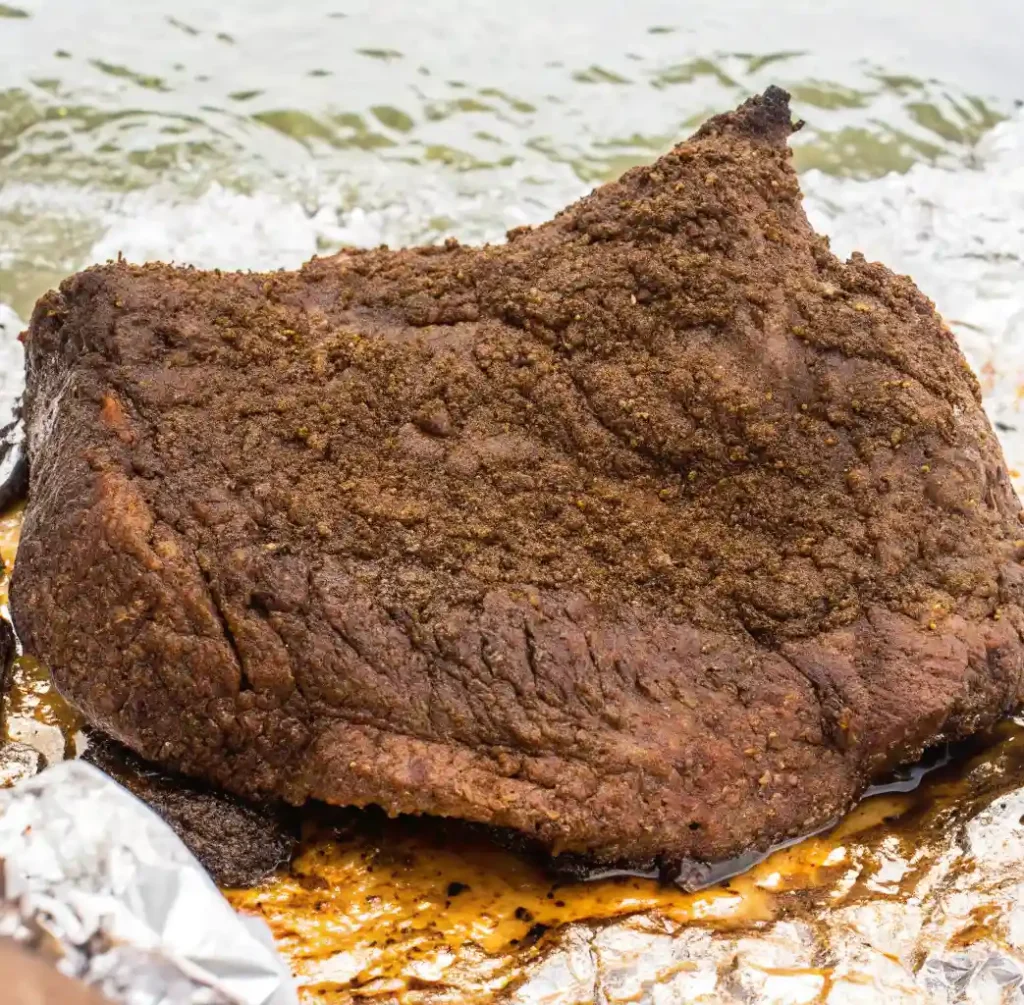 A whole smoked beef brisket with dark, thick seasoning, covered in aluminum foil with some of the juices extracted from the meat, revealing its delicious appearance and moist edges.