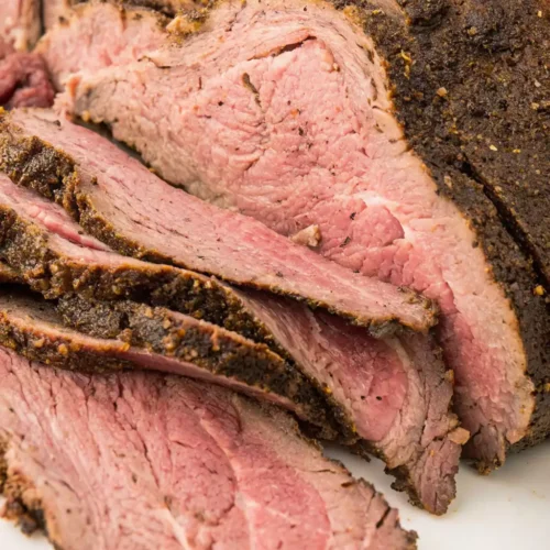 A close-up of a partially sliced smoked corned beef brisket shows a tender, rosy-pink interior and a dark, peppery crust, with the slices fanned out on a white serving surface.