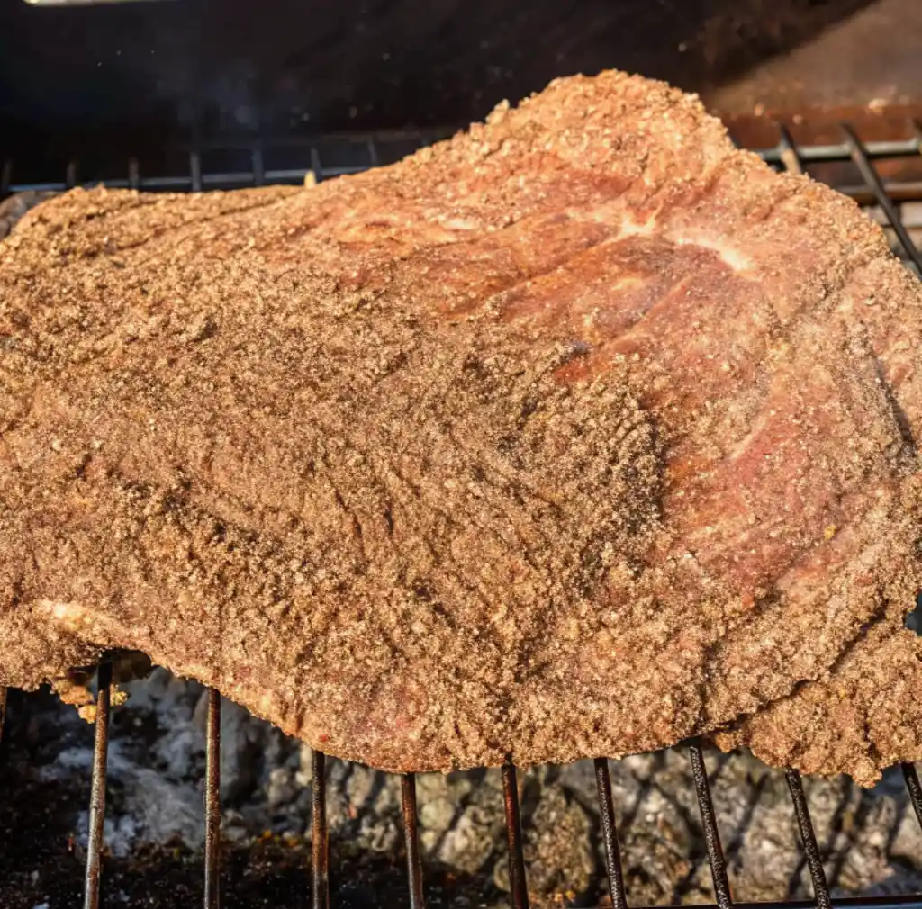 A corned beef brisket, heavily coated with a coarse, light brown spice rub, is shown resting directly on a dark wire grill grate over glowing embers and hot coals.