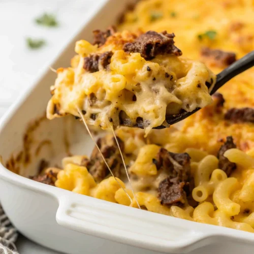 Brisket Mac and Cheese casserole in a white baking dish. A black spoon scoops out a creamy portion, showcasing the elbow macaroni, chunks of brisket, and a golden, bubbly baked cheese topping.