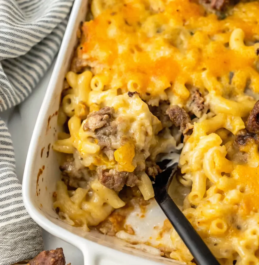 Brisket Mac and Cheese casserole in a white baking dish. A black spoon scoops out a creamy portion, showcasing the elbow macaroni, chunks of brisket, and a golden, bubbly baked cheese topping.
