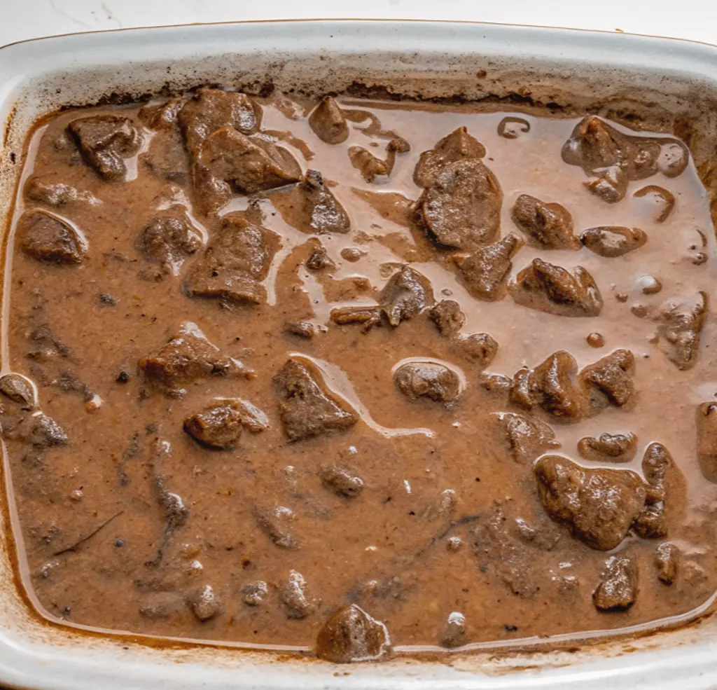 Beef cubes in a rich, thick brown gravy, filling a white rectangular baking dish. The tips are stewed to a dark, savory color