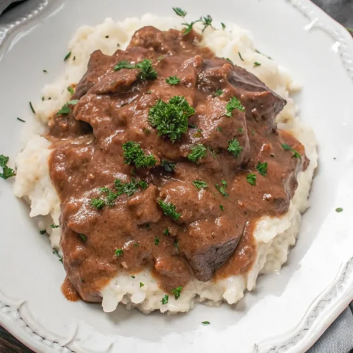 No Peek Beef Tips Recipe A large plate of tender, dark brown beef tips and thick gravy is placed on a mound of fluffy white mashed potatoes, garnished with fresh parsley on a white plate.