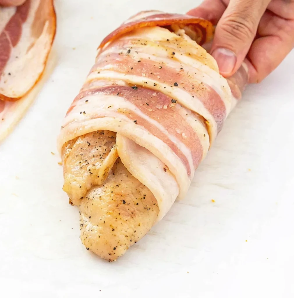 A hand is wrapping a seasoned chicken breast tightly with overlapping strips of uncooked bacon, preparing the ranch bacon-wrapped chicken for baking or grilling.