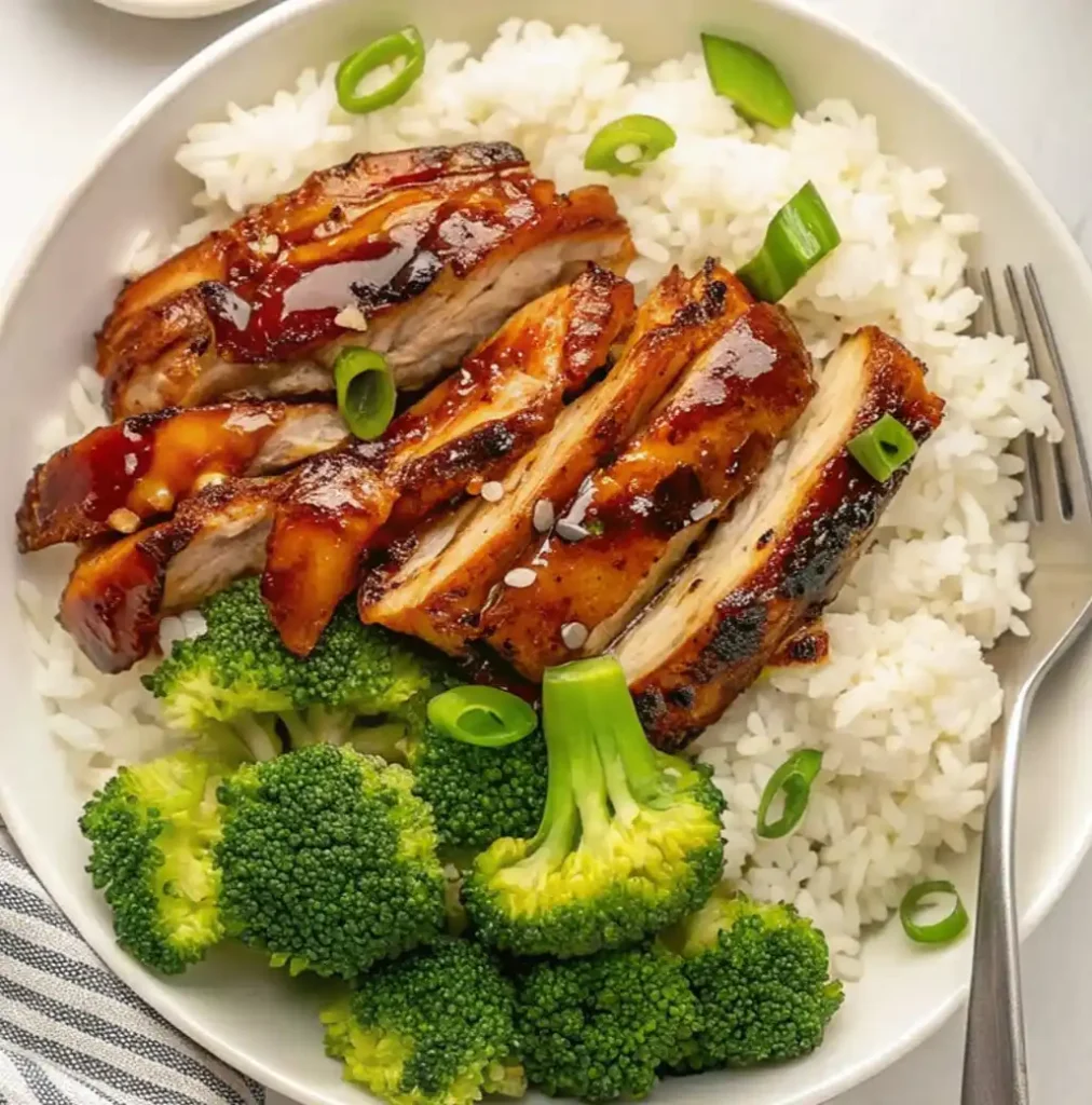 A white bowl filled with white rice, topped with slices of glossy, panda express grilled teriyaki chicken, chopped green onions, and a side of steamed broccoli florets.