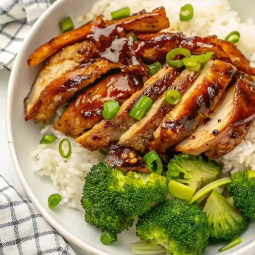 A white bowl filled with white rice, topped with slices of glossy, panda express grilled teriyaki chicken, chopped green onions, and a side of steamed broccoli florets.