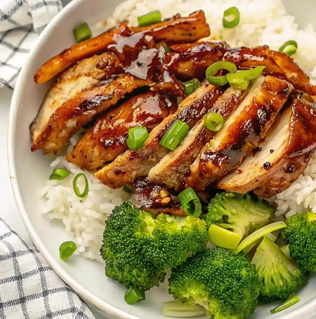 A white bowl filled with white rice, topped with slices of glossy, panda express grilled teriyaki chicken, chopped green onions, and a side of steamed broccoli florets.