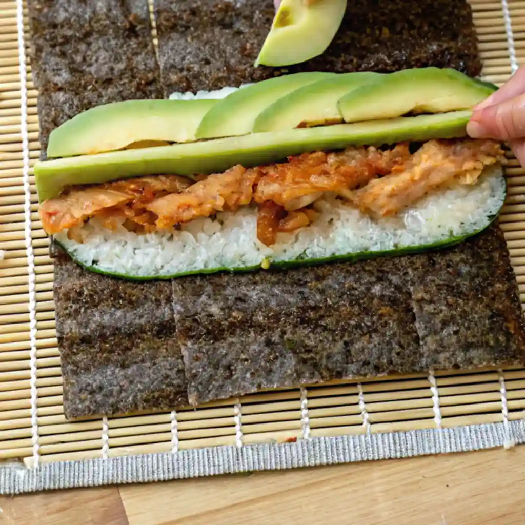 Assembling a sushi roll with rice, tempura chicken, cucumber, and avocado on a nori sheet and bamboo mat.