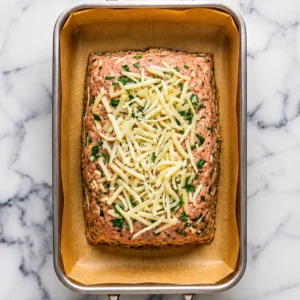 unbaked chicken cordon bleu meatloaf shaped in a parchment-lined baking pan, topped with a generous layer of shredded Swiss cheese and chopped herbs.