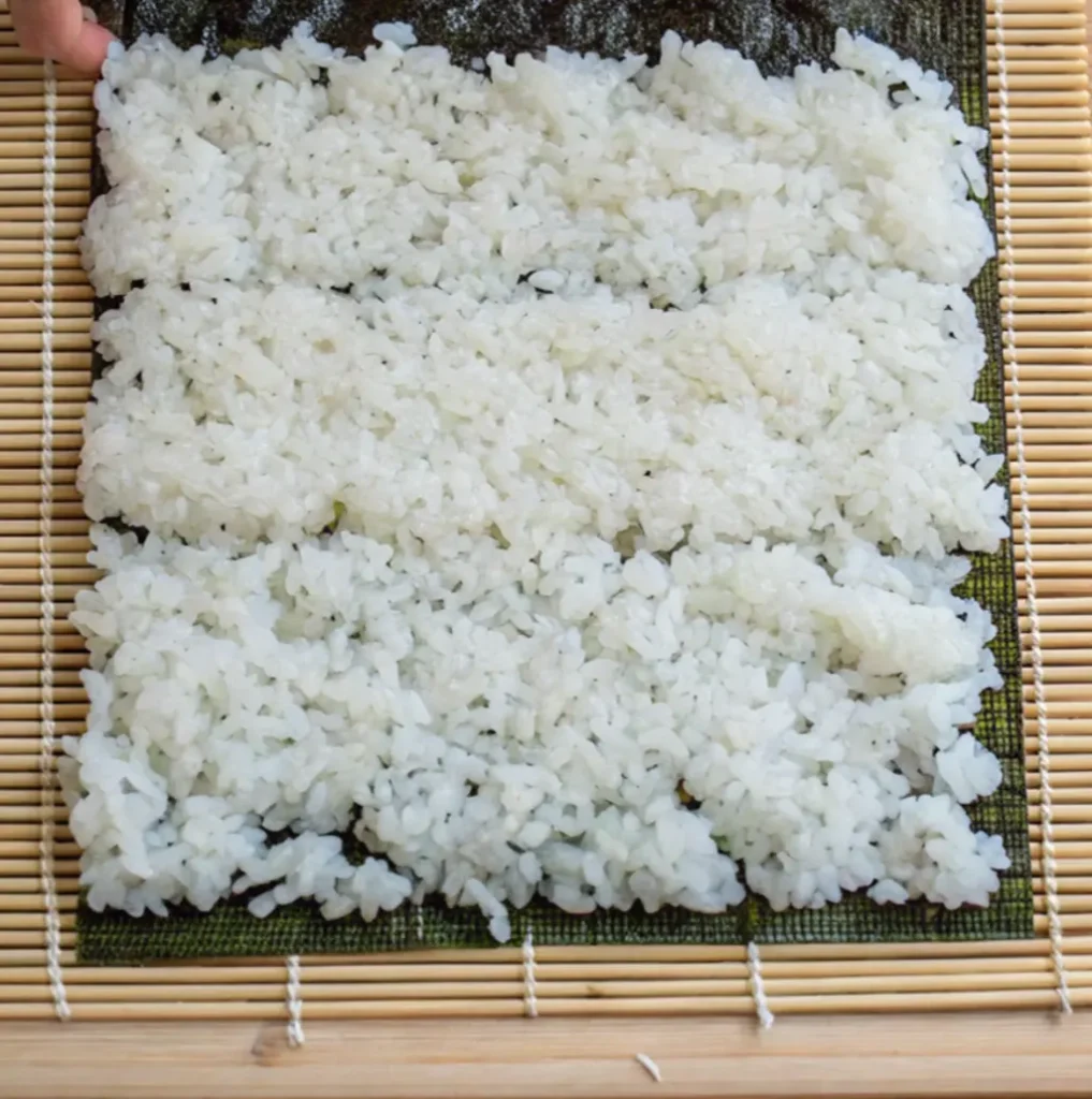 A rectangular layer of white, sticky sushi rice spread evenly over a sheet of dark green nori (seaweed) on a bamboo rolling mat, preparing for a sushi roll.