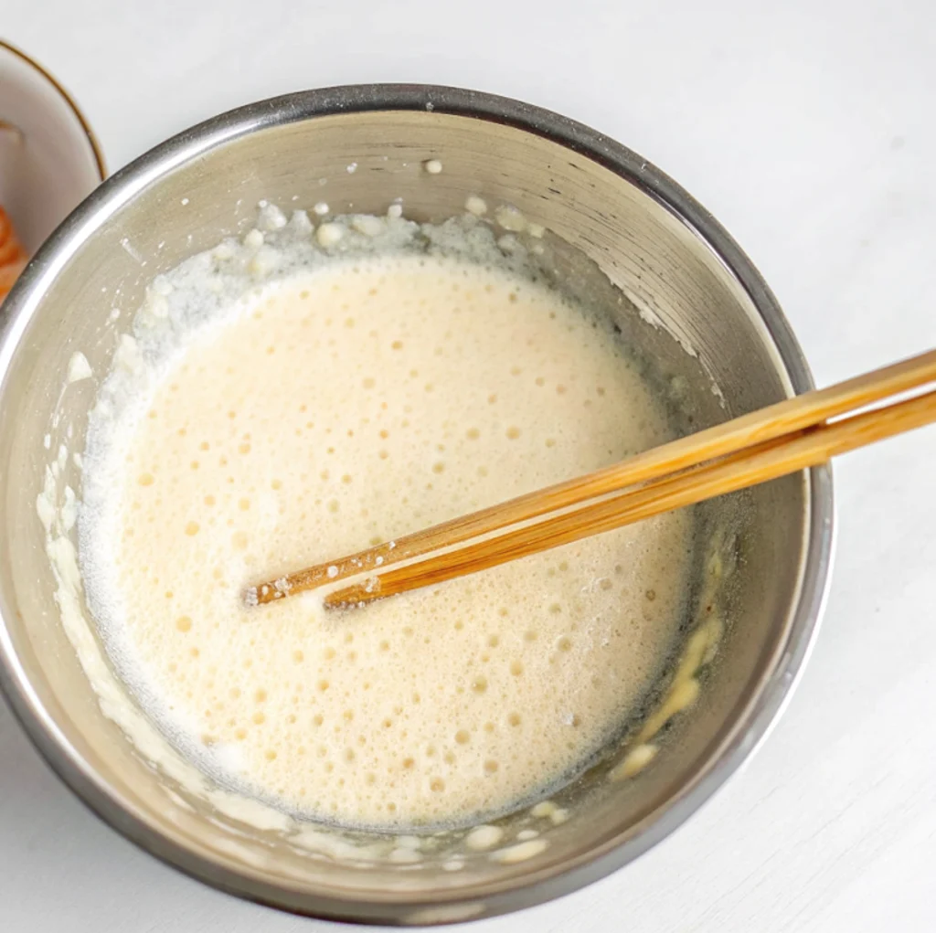 smooth, bubbly tempura batter mix in a metal bowl, stirred slightly by a pair of bamboo chopsticks on a