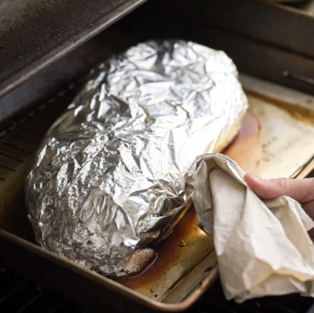 The partially cooked beef brisket is tightly wrapped in aluminum foil during the smoking stage, with one hand holding a piece of cloth.