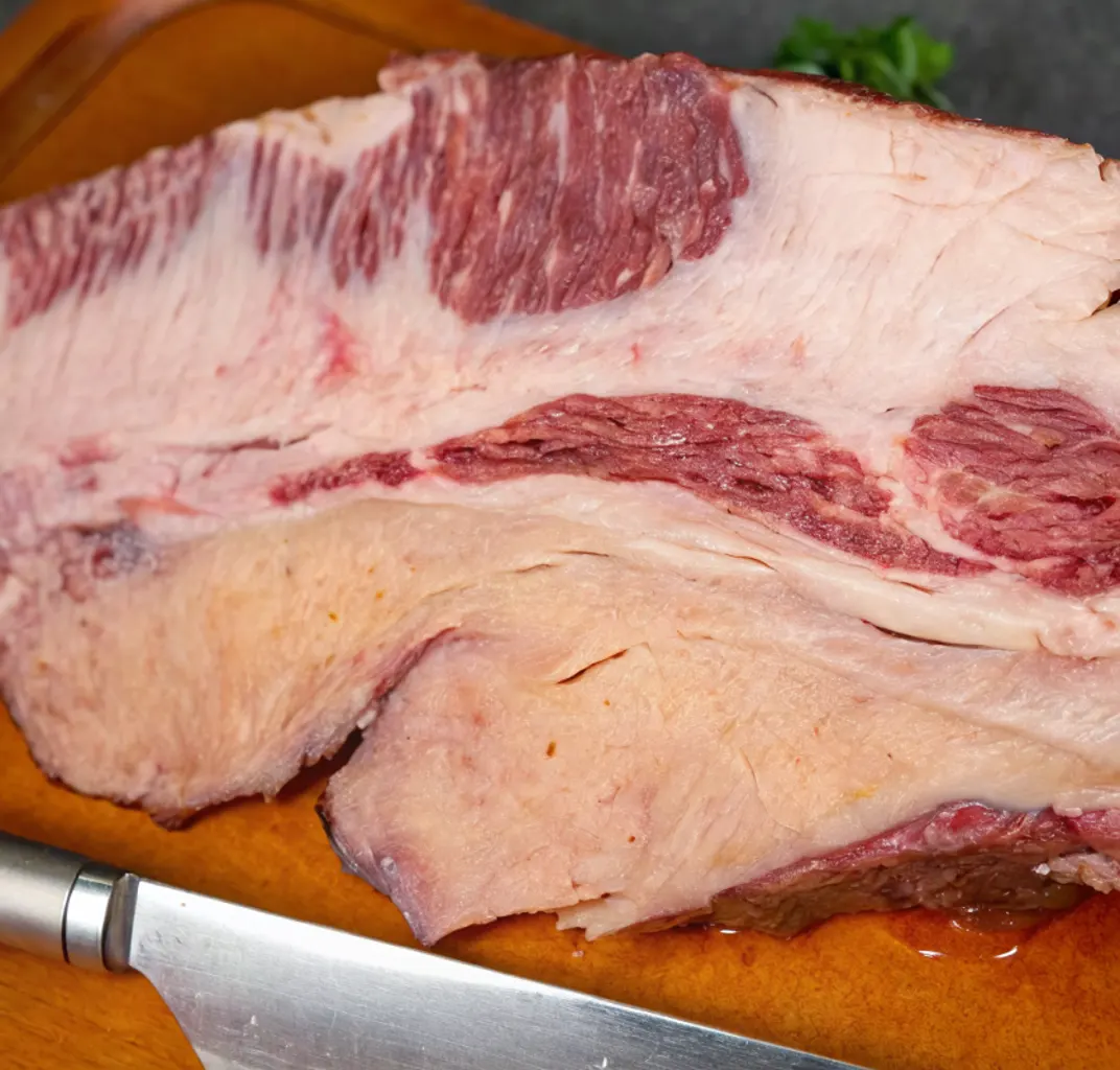 An uncooked beef brisket sitting on a wooden cutting board with a silver knife resting beneath it.