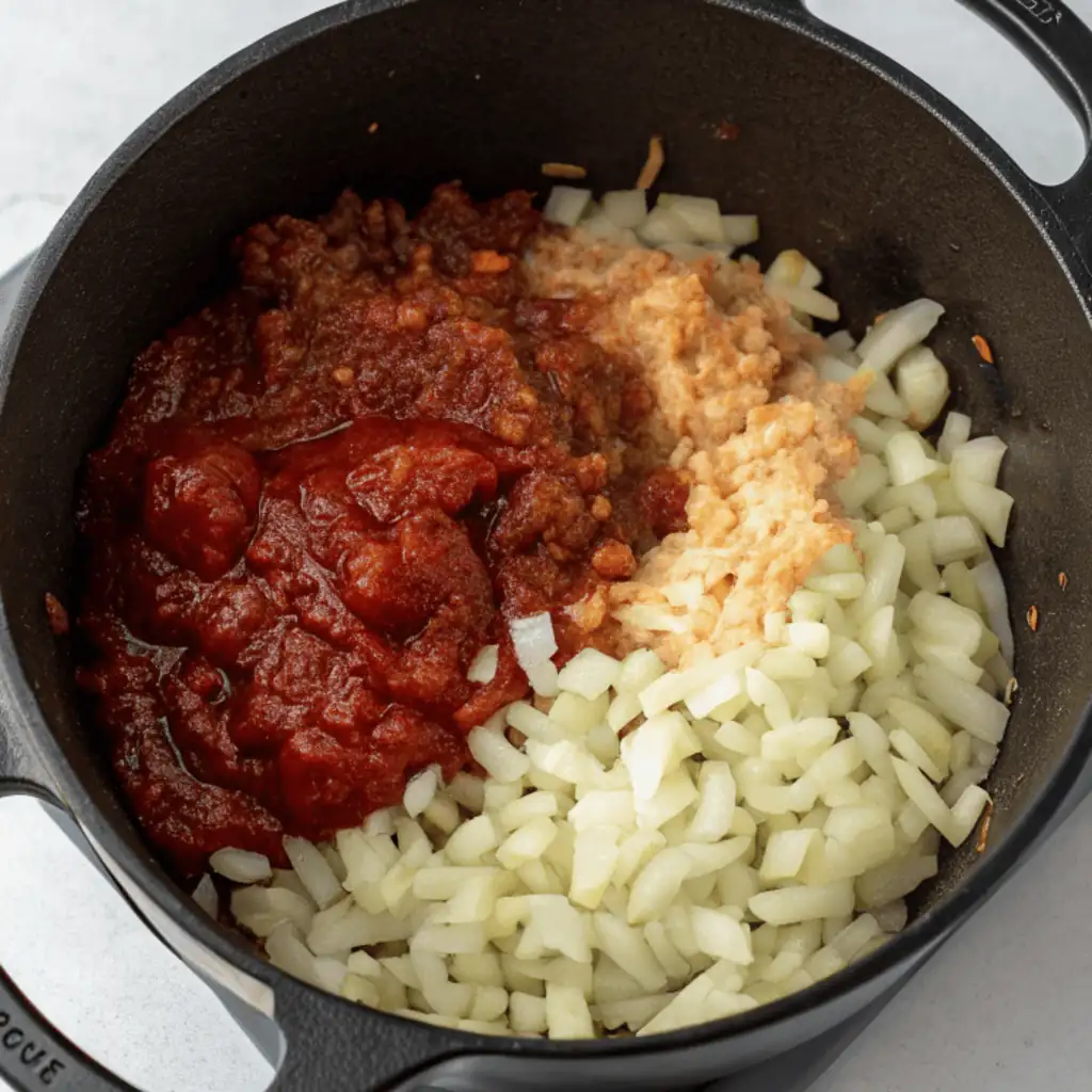 A frying pan on a stovetop with chopped onions, a dark red paste, and a lighter brown mixture.