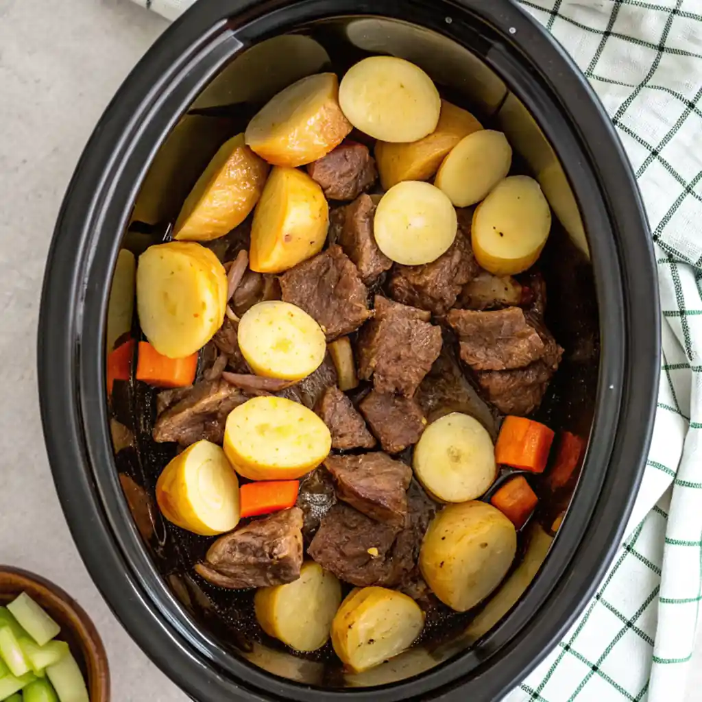 Sliced meat, potatoes and vegetables placed in a black slow cooker and ready to cook for hours.