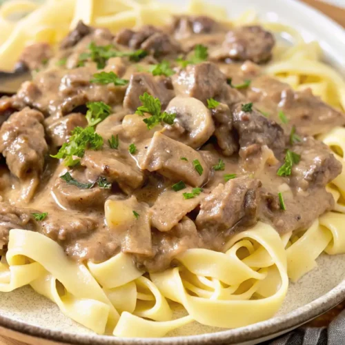 serving of Gluten Free Beef Stroganoff. Tender beef and sliced mushrooms are coated in a rich, creamy, brown sauce and served over a bed of wide, flat gluten-free noodles. It is garnished with fresh parsley.