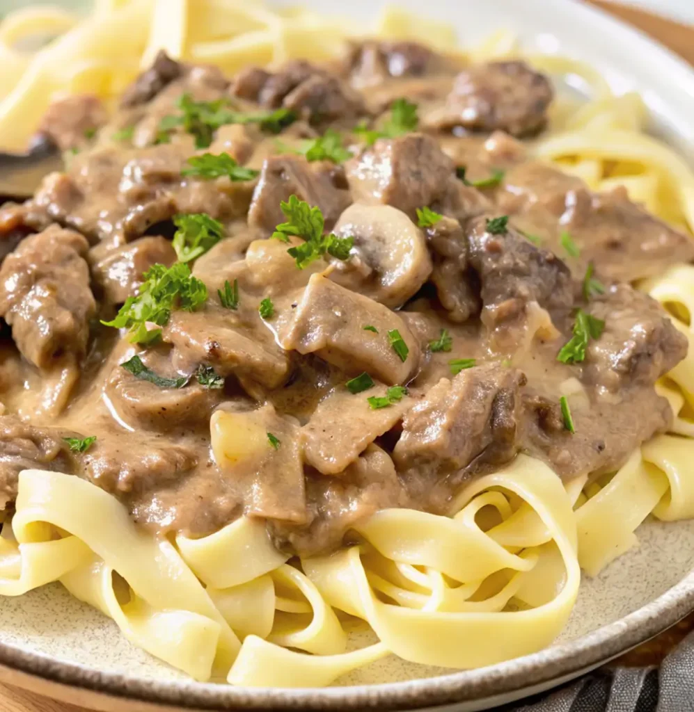 serving of Gluten Free Beef Stroganoff. Tender beef and sliced mushrooms are coated in a rich, creamy, brown sauce and served over a bed of wide, flat gluten-free noodles. It is garnished with fresh parsley.