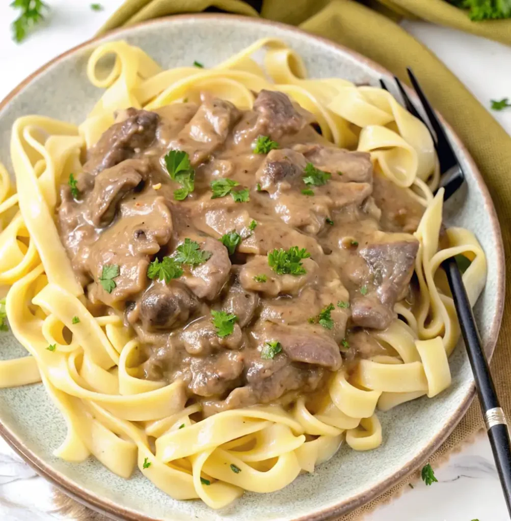 serving of Gluten Free Beef Stroganoff. Tender beef and sliced mushrooms are coated in a rich, creamy, brown sauce and served over a bed of wide, flat gluten-free noodles. It is garnished with fresh parsley.