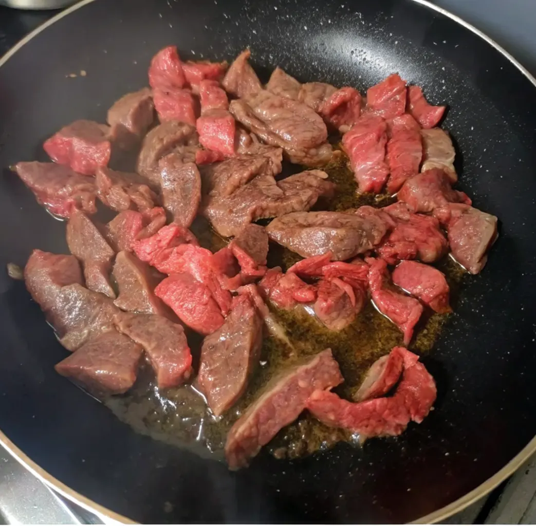 strips of beef being seared in a dark non-stick pan with a small amount of oil. Some pieces are already browning, while others are still a vibrant red
