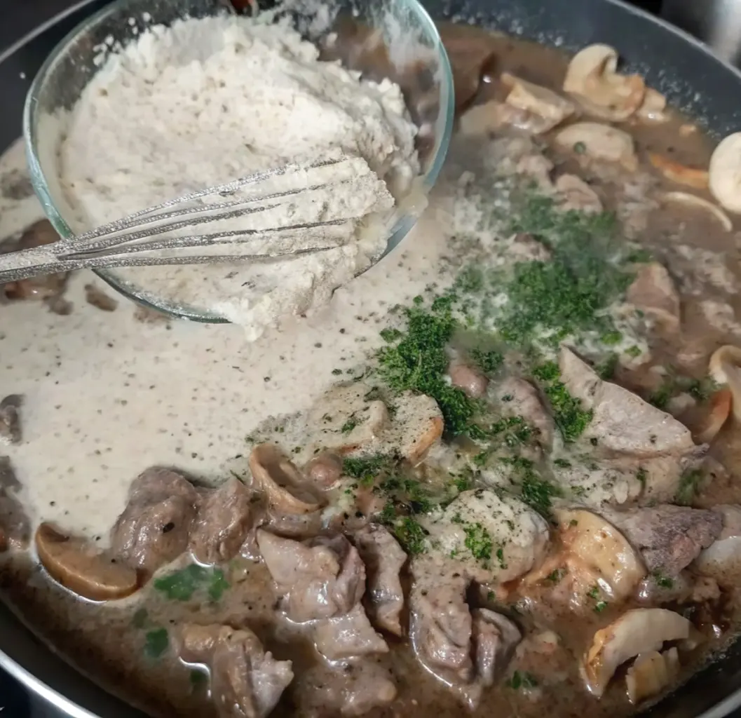Thicken and enrich the stroganoff sauce with beef. Pour a bowl of gluten-free flour into the pan over the cooked beef and mushrooms, which have been seasoned with parsley.