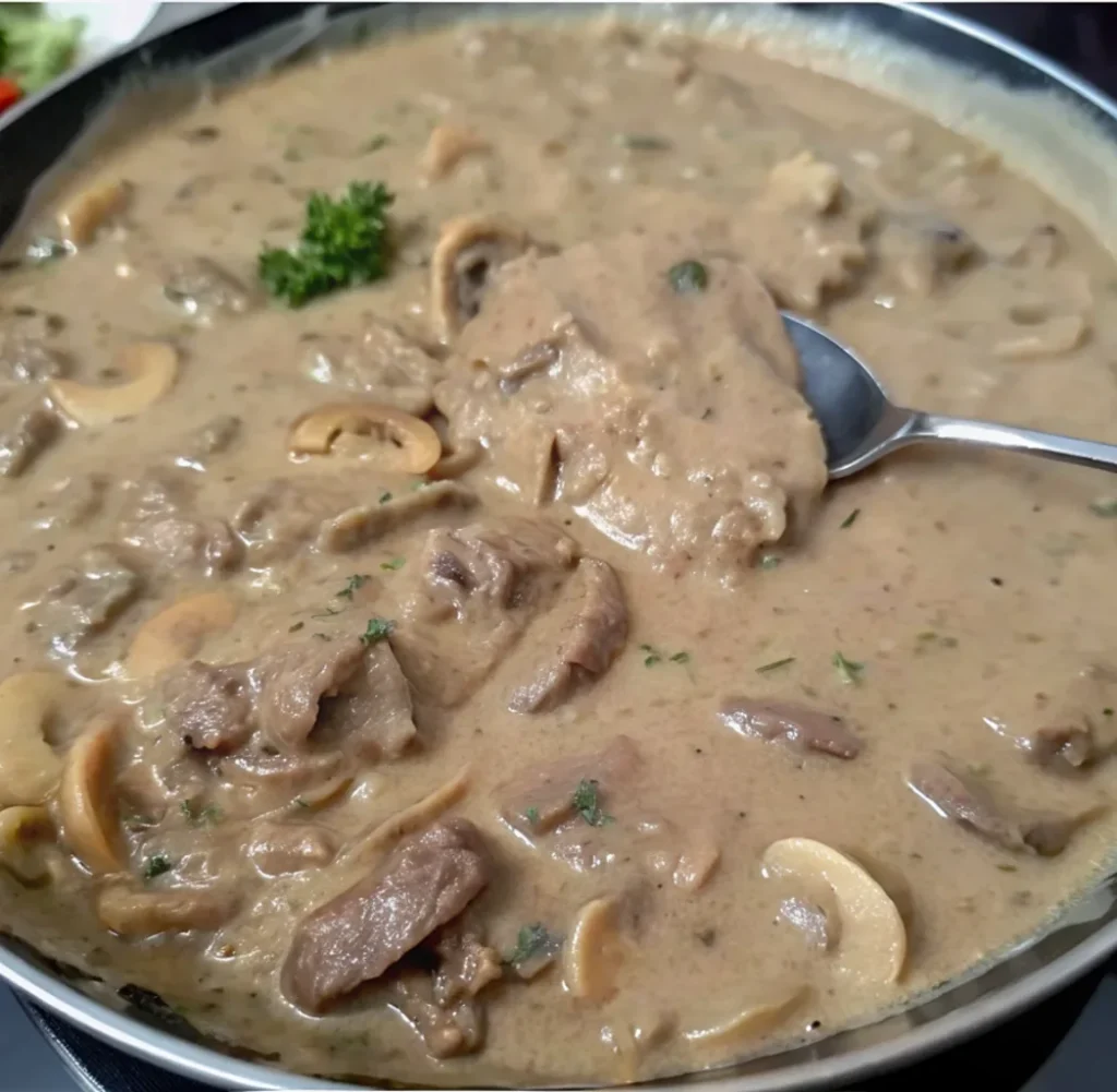 the finished Gluten-Free Beef Stroganoff simmering in a pan. The tender beef and sliced mushrooms are completely coated in the thick, rich, and creamy brown sauce. A spoonful lifts some of the mixture to show its texture.